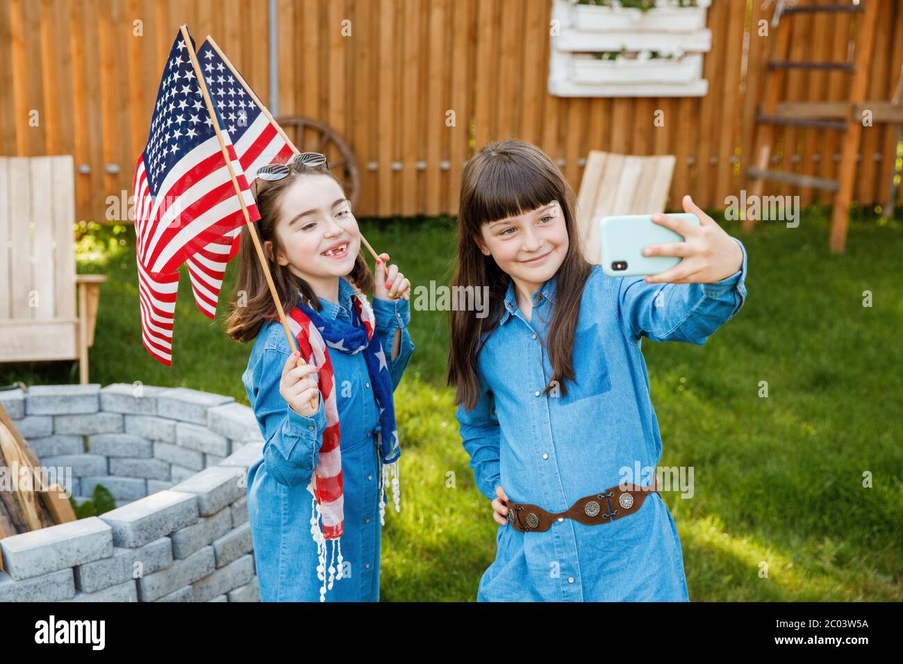 Zwei Mädchen stehen auf dem Hof, halten die Flagge der USA und fotografieren sich auf einem Handy Stockfoto