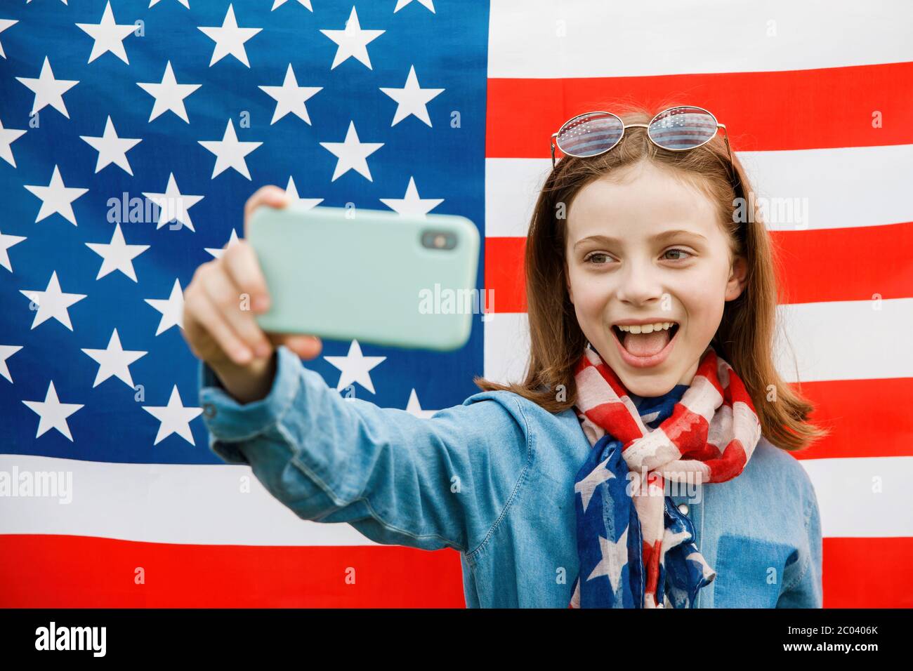 Fröhliche hübsche Mädchen steht auf dem Hintergrund der amerikanischen Flagge und macht Bilder von sich auf einem Handy Stockfoto