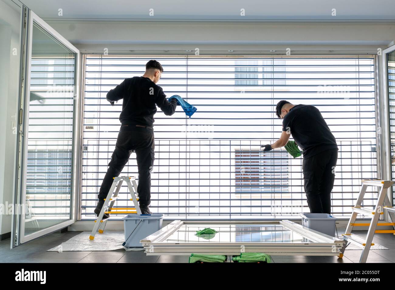 Horizontale Ansicht von zwei männlichen professionellen Reinigern Reinigung Jalousien auf einer großen Fensterfront mit Mikrofasertüchern Stockfoto