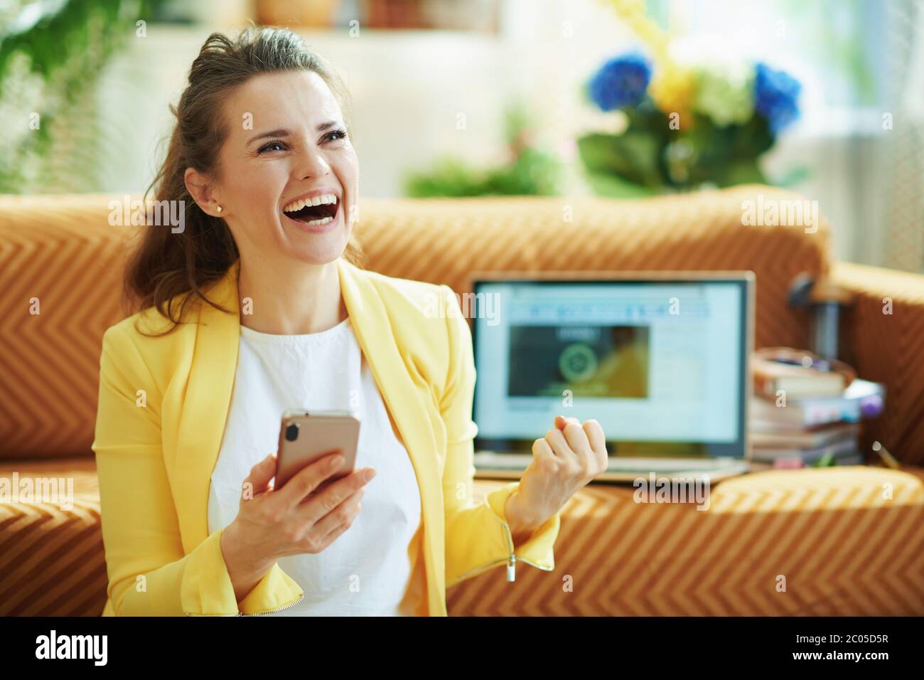 Glücklich elegante Mittelalterfrau in gelber Jacke mit Laptop und Smartphone jubeln während des Studiums online im modernen Haus an sonnigen Tag. Stockfoto Glücklich elegante Mittelalterfrau in gelber Jacke mit Laptop und Smartphone jubeln während des Studiums online im modernen Haus an sonnigen Tag. Stockfoto