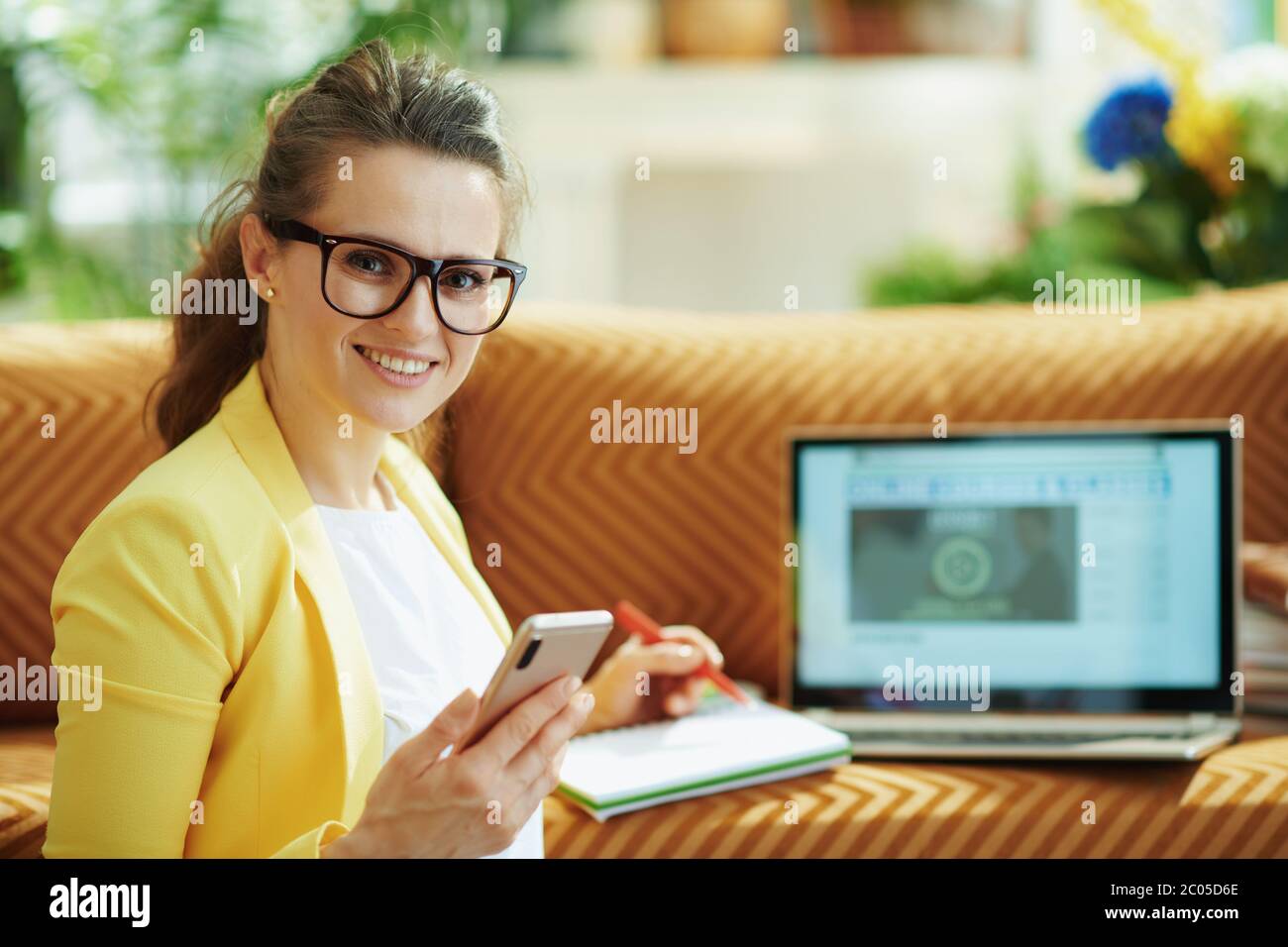 Portrait der glücklichen jungen Frau in gelber Jacke mit Laptop und Smartphone Online studieren im modernen Wohnzimmer an sonnigen Tag. Stockfoto Portrait der glücklichen jungen Frau in gelber Jacke mit Laptop und Smartphone Online studieren im modernen Wohnzimmer an sonnigen Tag. Stockfoto
