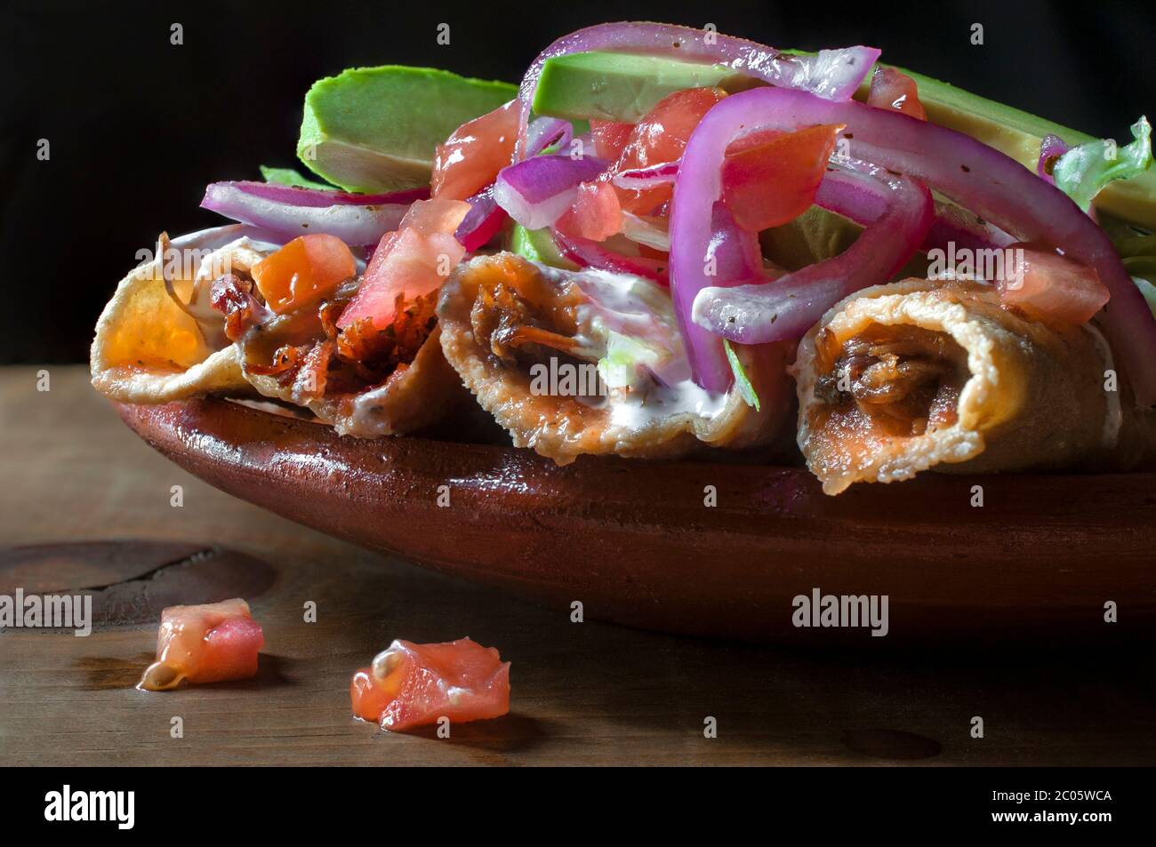 Tacos dorados auf einem mexikanischen Lehm oder Schlammteller, mit lila Zwiebeln, Avocado, Tomatencreme und würziger Salsa. Mexikanisches Essen auf schwarzem Hintergrund. Stockfoto