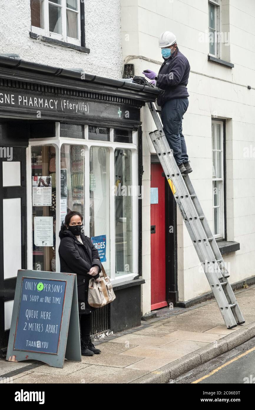 Ein Telefoningenieur, der einen Hut, eine Gesichtsmaske und Handschuhe trägt, während er außerhalb einer Apotheke arbeitet, neben einem ähnlich maskierten Kunden, Presteigne, Wales, Großbritannien Stockfoto