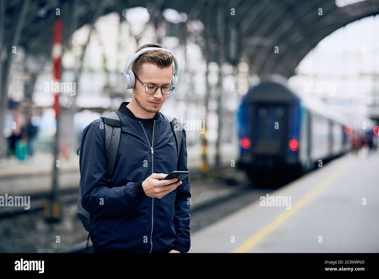 Junger Mann mit Kopfhörern, der Musik am Bahnhof mit dem Telefon gegen den Zug hört. Stockfoto