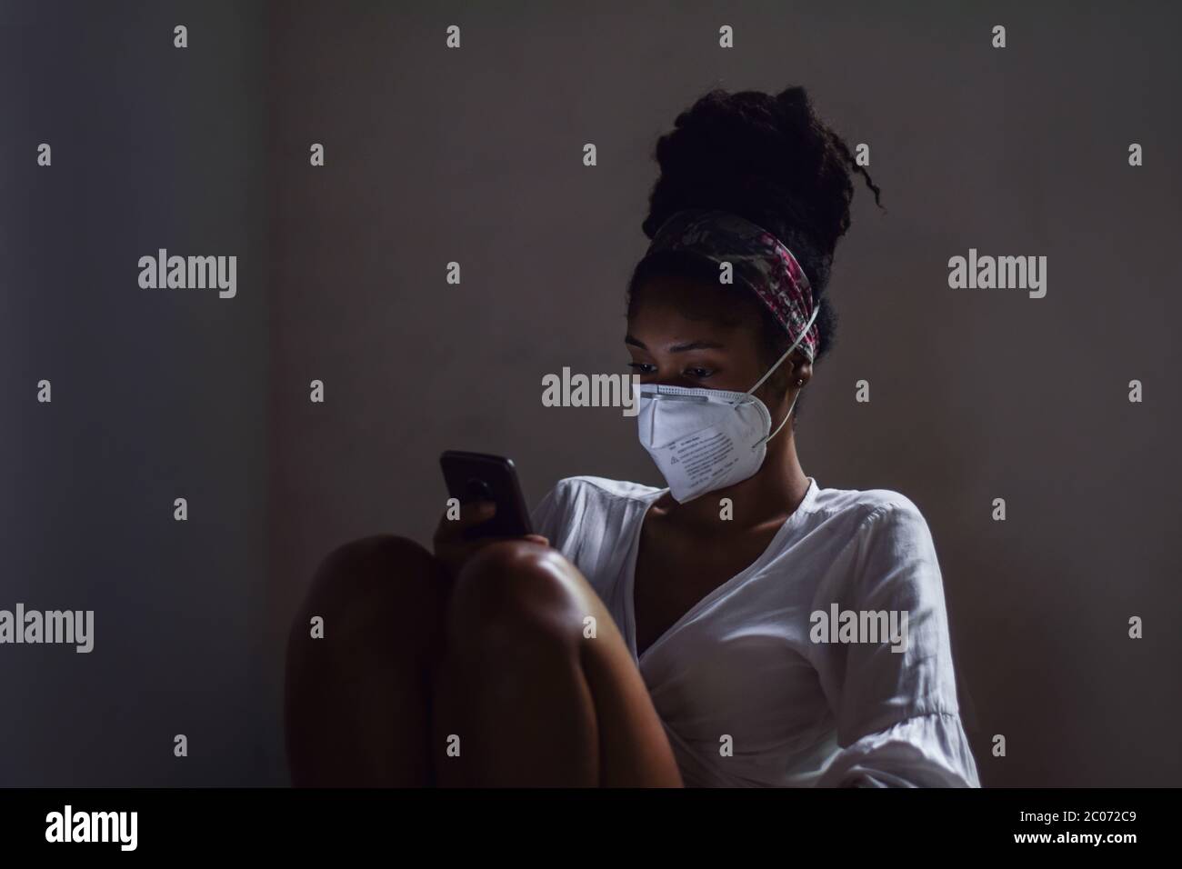 Junge Frau in Gesichtsmaske mit Smartphone im dunklen Raum Stockfoto