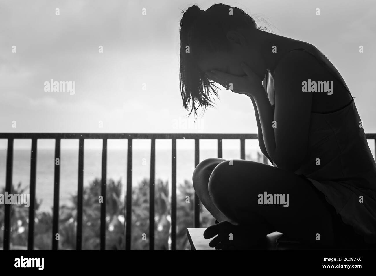 Silloutte einer Frau, die allein auf ihrem Hotelbalkon sitzt und ihr Gesicht weint und schwarz und weiß bedeckt. Stockfoto