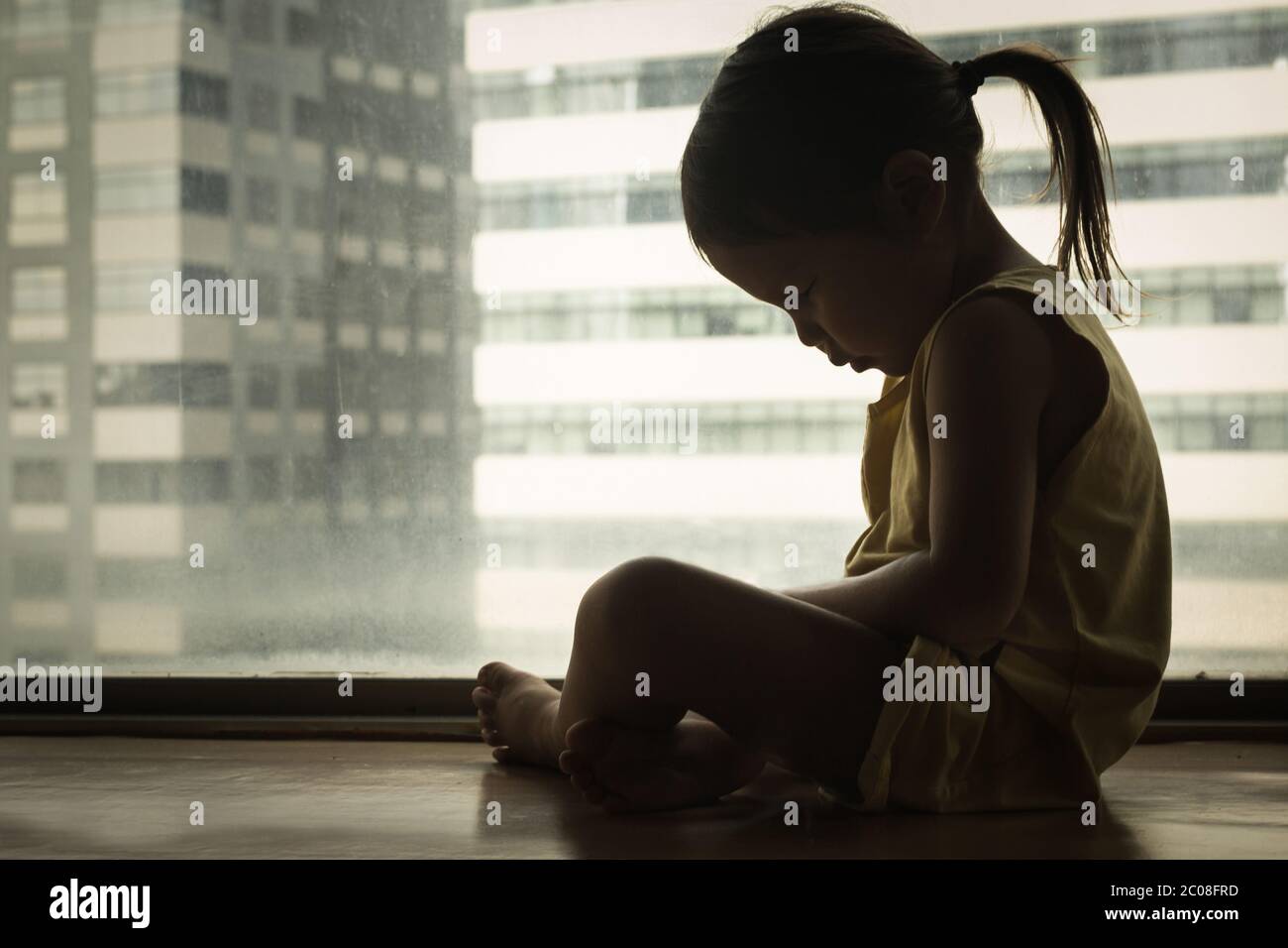 Ein kleines Mädchen, das neben einem Fenster sitzt und den Kopf in Traurigkeit nach unten drückt. Gefühl deprimiert und verletzt. Stockfoto