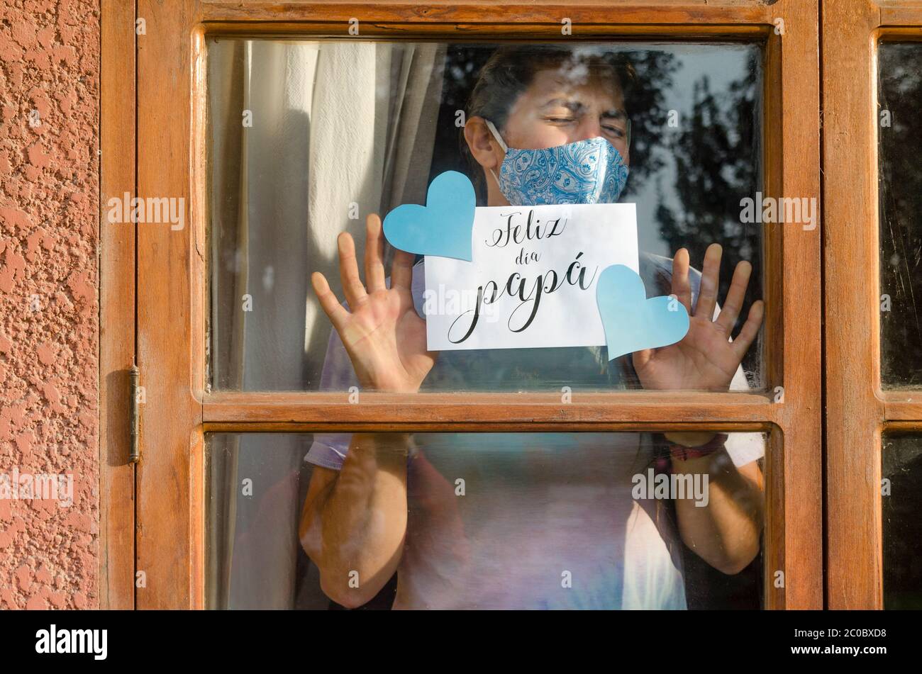 Junge Frau mit Gesichtsmaske zu Hause, die ein Blatt Papier durch das Fenster hält, das auf spanisch sagt: 'Glücklicher Tag, Papa!' Stockfoto