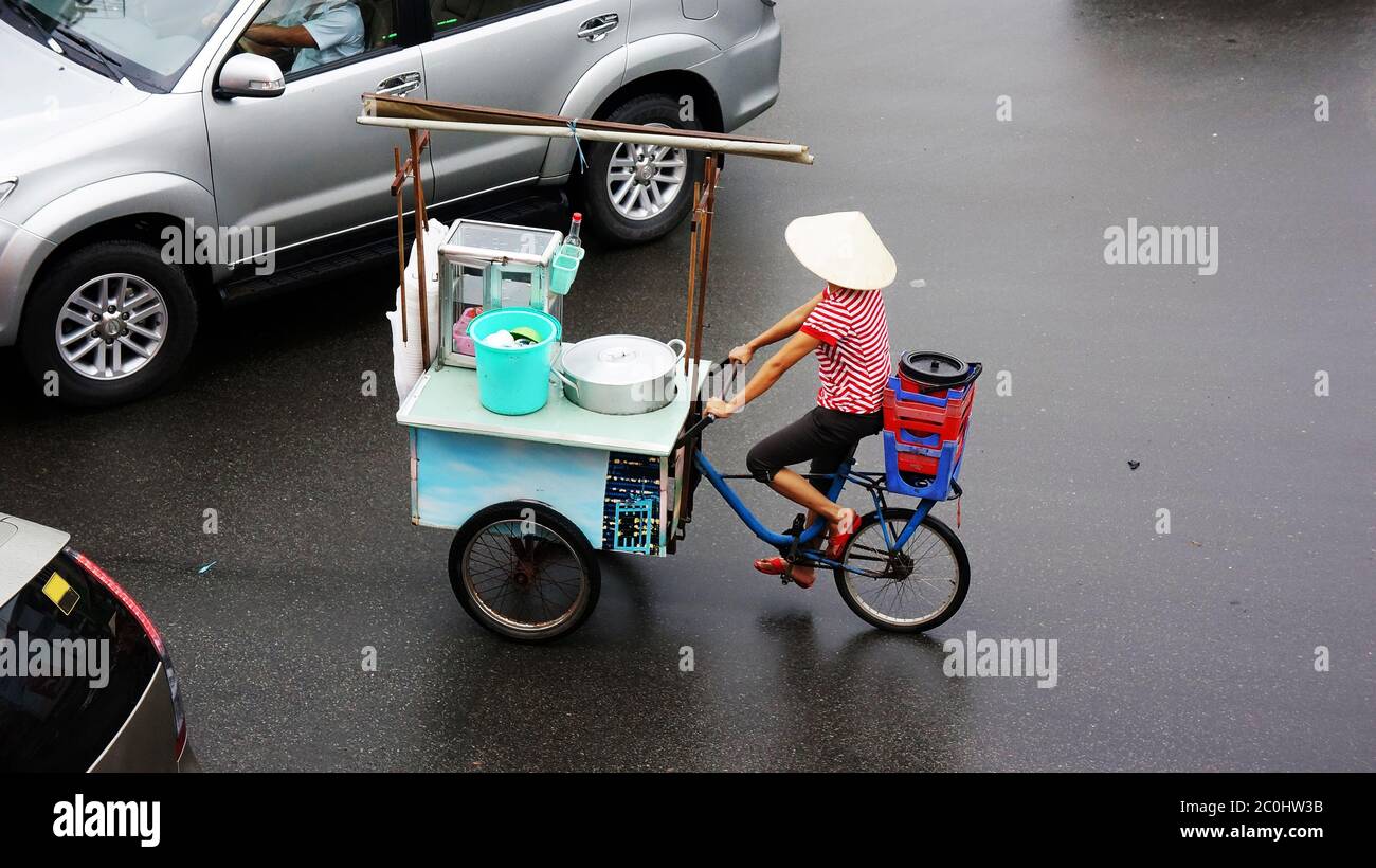 Vietnamesische Frau fahren drei Rad Fahrrad auf nasser Straße durch Regen und Transfer mobile Restaurant des Lebensmittelverkäufers als Kunststoff-Hocker, großen Topf Stockfoto