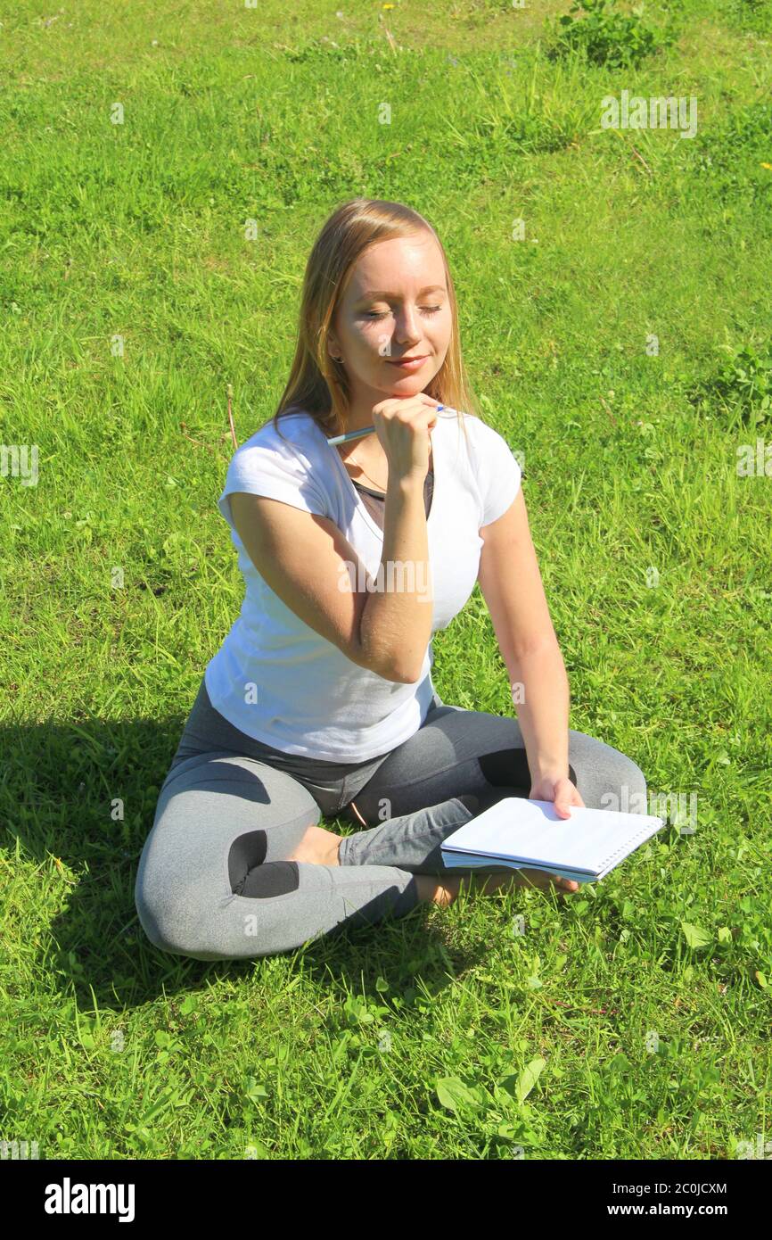 Ein junges, schönes Mädchen im weißen T-Shirt mit geschlossenen Augen sitzt auf dem grünen Gras auf dem Rasen mit einem Notizbuch und einem Stift, mit dem Handkinn gestützt, denkt und träumt, komponiert Text und Gedichte. Stockfoto