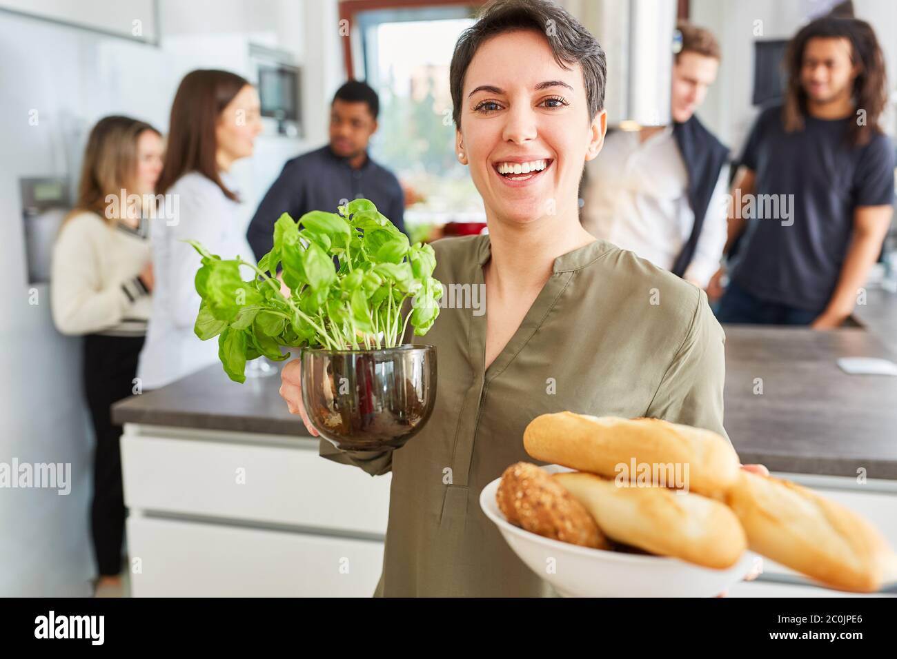 Frau mit Baguette und Basilikum in einer Gemeinschaftsküche mit Freunden im Hintergrund Stockfoto