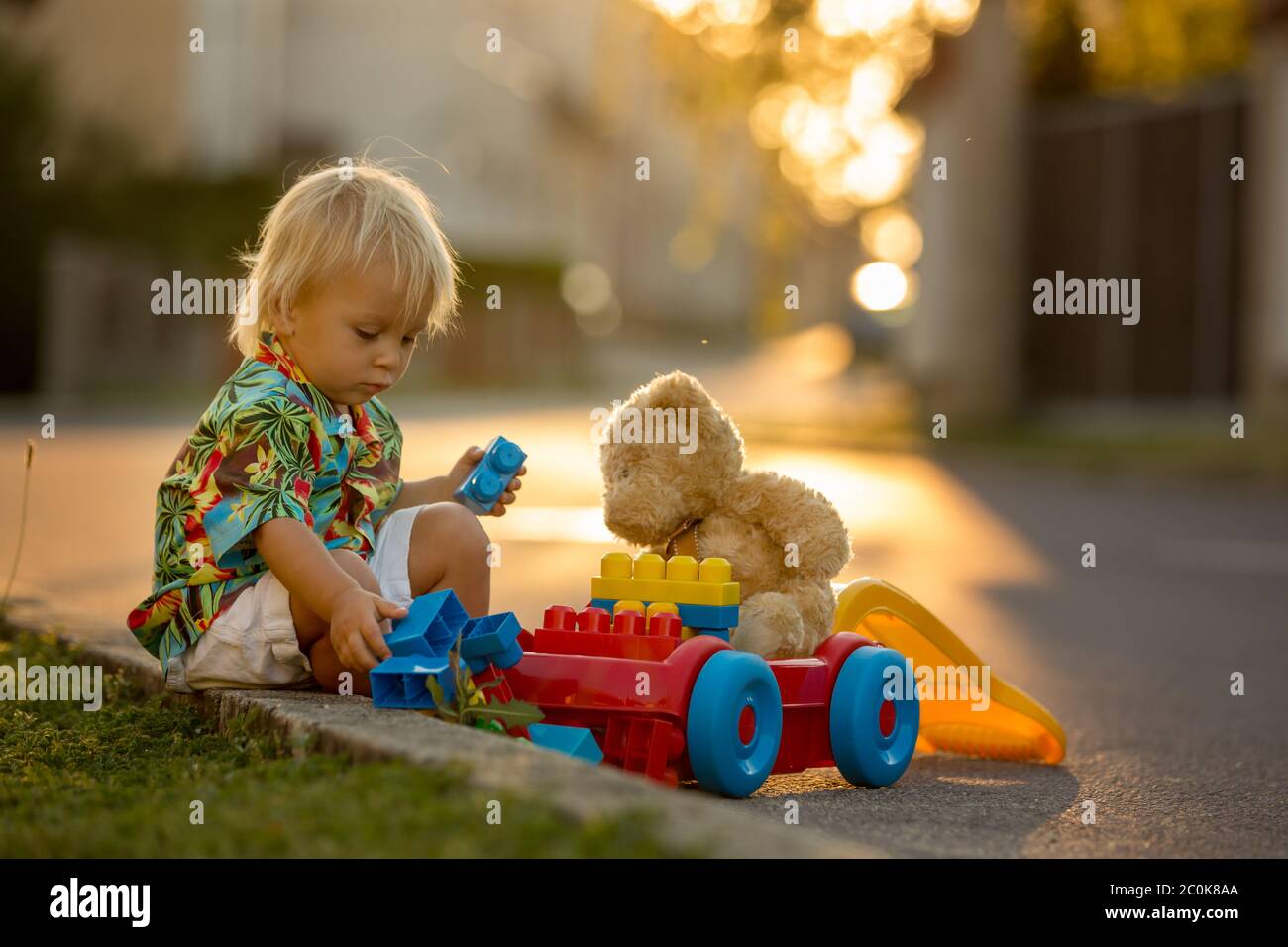 Schönes Kleinkind Kind, das mit Plastikspielzeug, Blöcken, Autos auf Sonnenuntergang in einem kleinen Dorf spielt Stockfoto