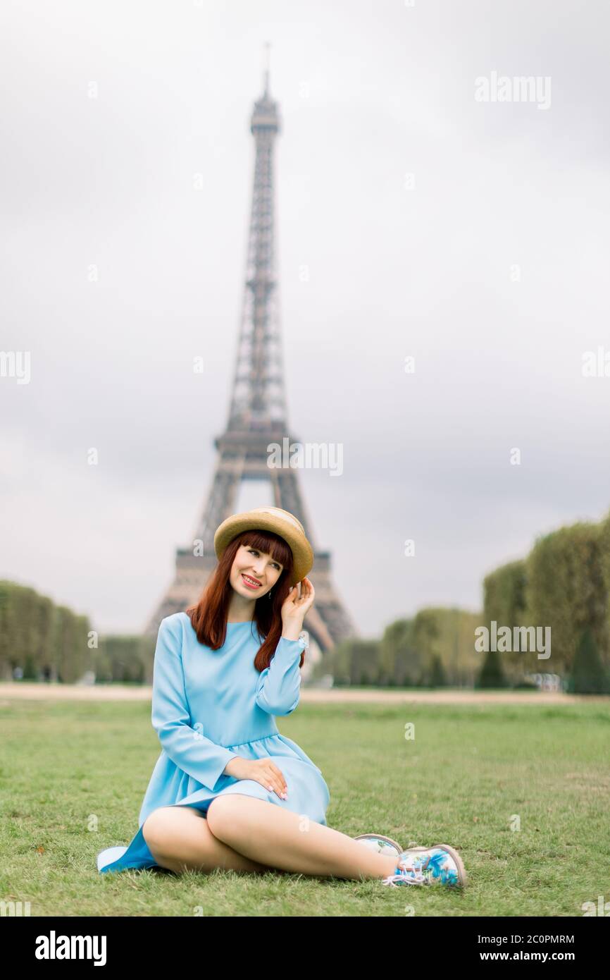 Elegante Frau in Strohhut und blauem Kleid sitzt auf grünem Gras in einem Park mit Eiffelturm auf dem Hintergrund Stockfoto