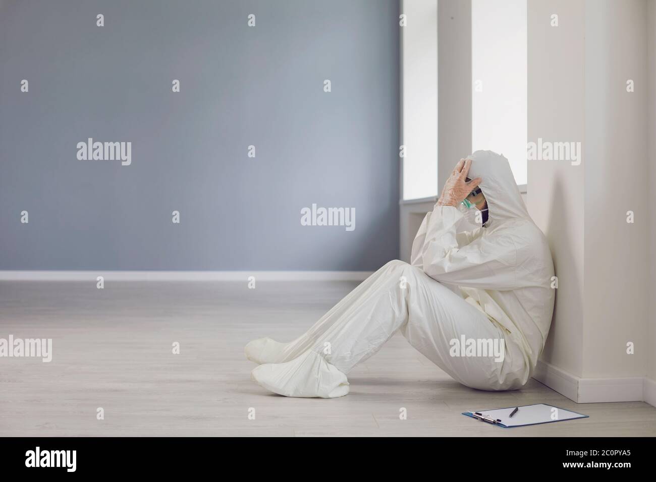 Verzweifelter Arzt in Schutzkleidung sitzt auf dem Boden in der Krankenhaushalle. Hoffnungsloser Sanitäter in Hazmat Anzug in der Klinik Stockfoto