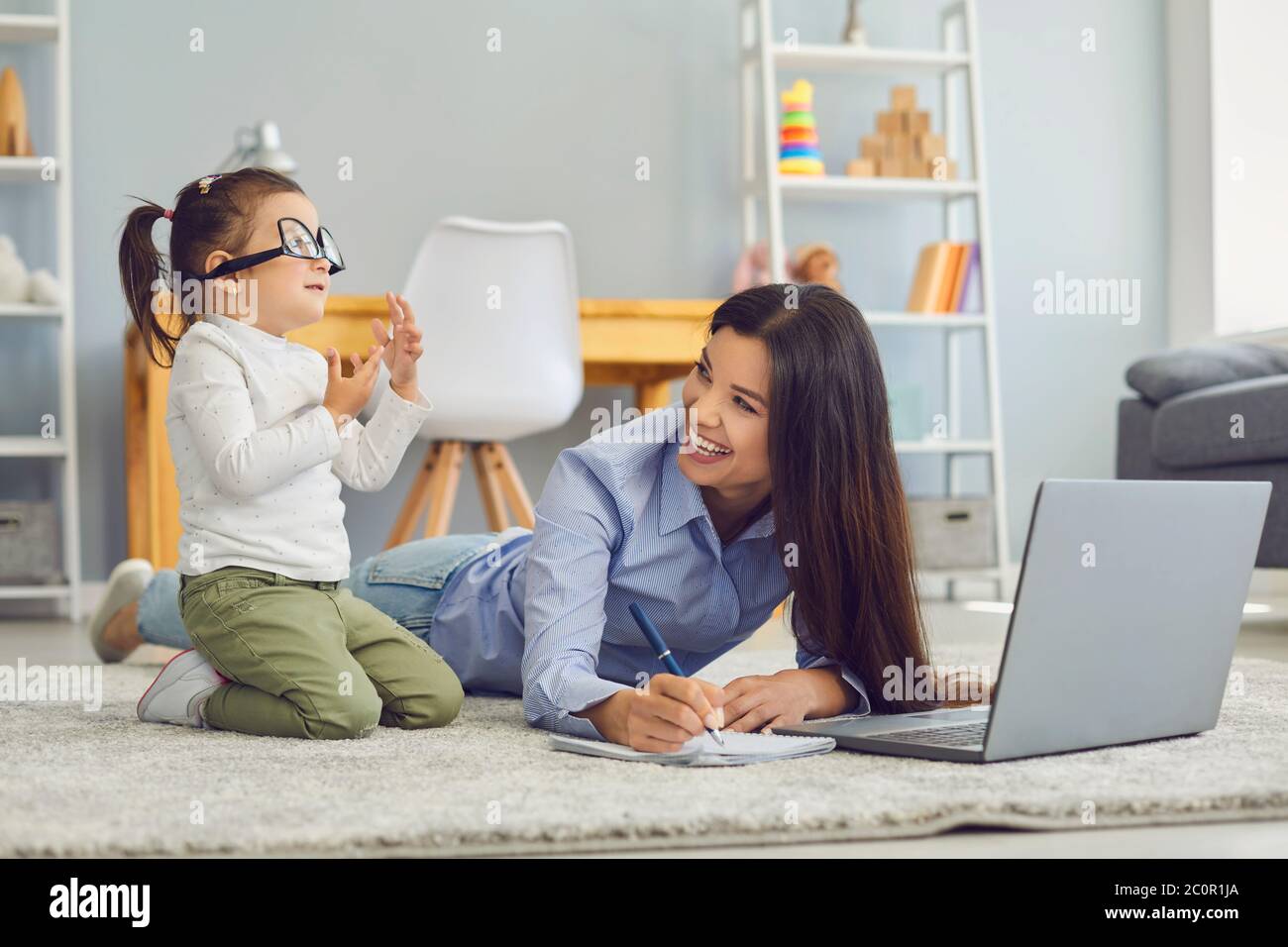 Junge Mutter arbeitet oder studiert auf Laptop, während ihre nette Tochter zu Hause alberne Spiele spielt. Multitasking und Life Balance Konzept Stockfoto