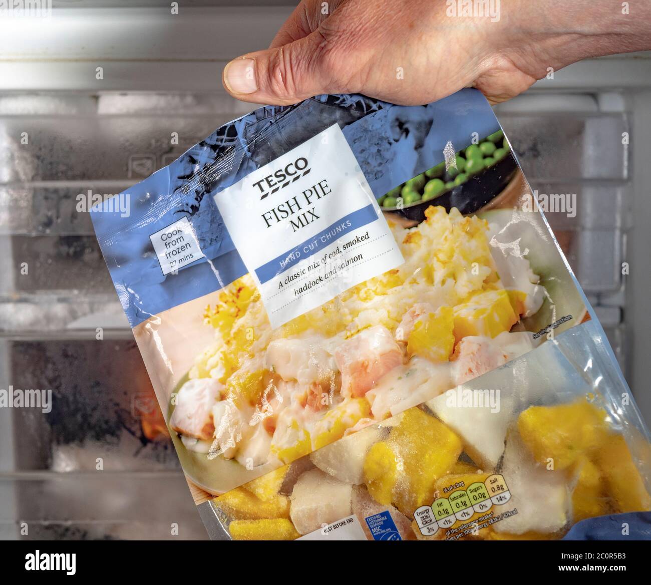 Eine Männerhand, die eine Tüte Tesco (eigener Hersteller) Fischpastete-Mischung – Kabeljau, geräucherter Schellfisch und Lachs – vor einem Gefrierschrank hält. Stockfoto