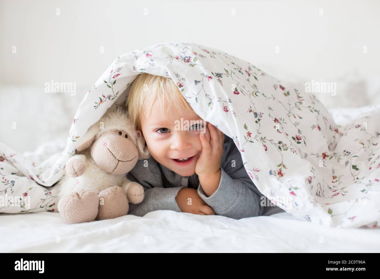 Kleiner kleiner kleiner Junge, der mit Teddyspielzeug spielt, sich unter der Decke im Bett versteckt, sonniges Schlafzimmer Stockfoto