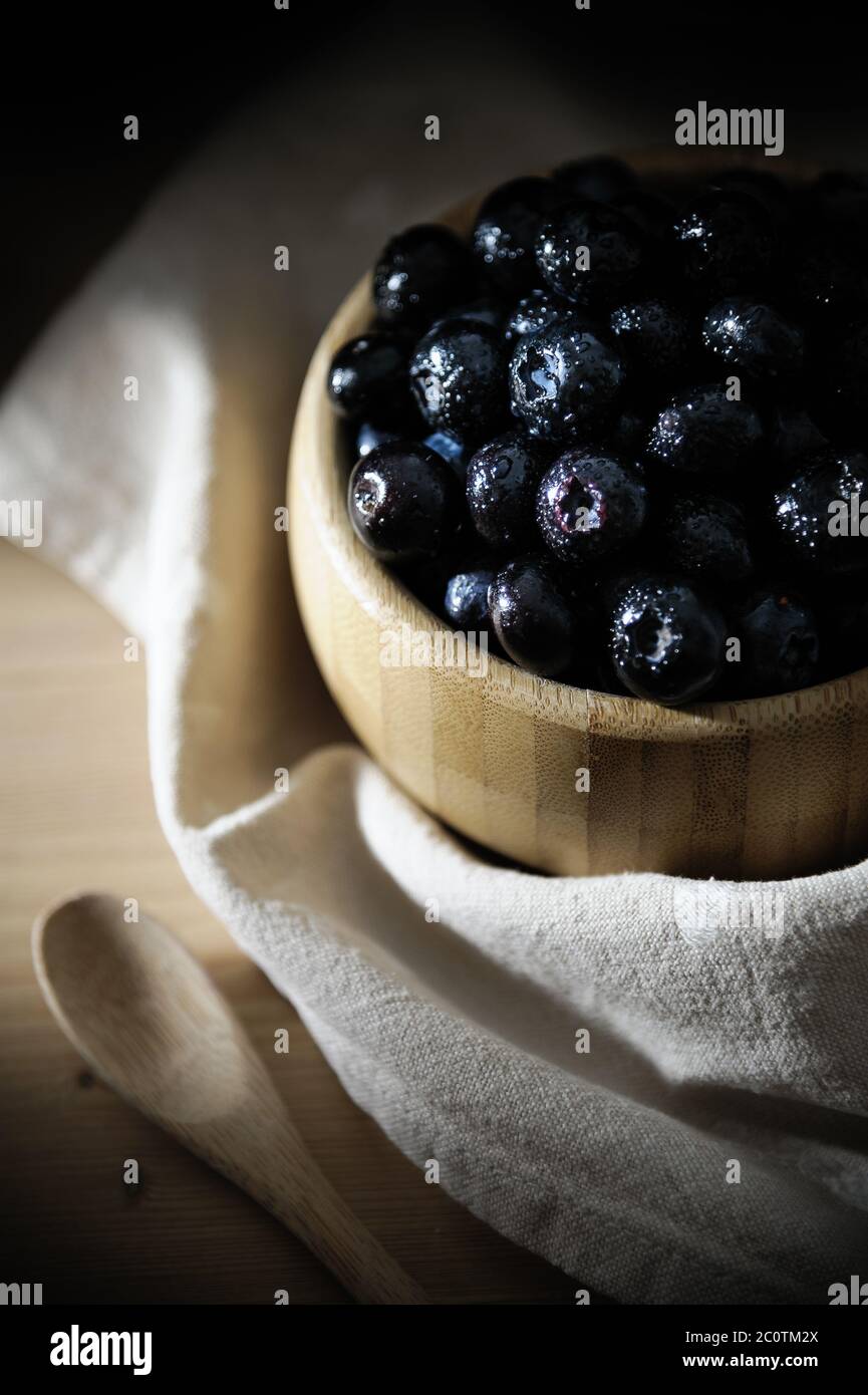 Frische Blaubeeren mit kleinen Tropfen Wasser auf der Oberfläche, in einer Bambusschale auf einem Holztisch, mit einer Tischdecke unten und einem Holzlöffel. Stockfoto