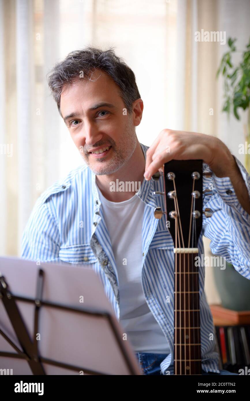 Nahaufnahme Porträt eines lächelnden Mannes, der sich auf dem Kopfteil einer Gitarre stützt und zu Hause Kamera und Musikständer mit Abschied vor dem Kopf anschaut. Stockfoto