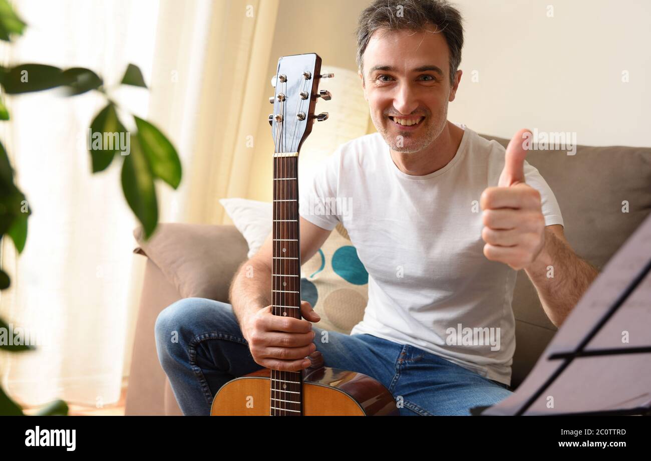 Lächelnder Mann mit seiner akustischen Gitarre in der Hand schauend Augen mit ok Geste sitzend auf dem Sofa mit Musik-Stand mit Songs zu Hause Stockfoto