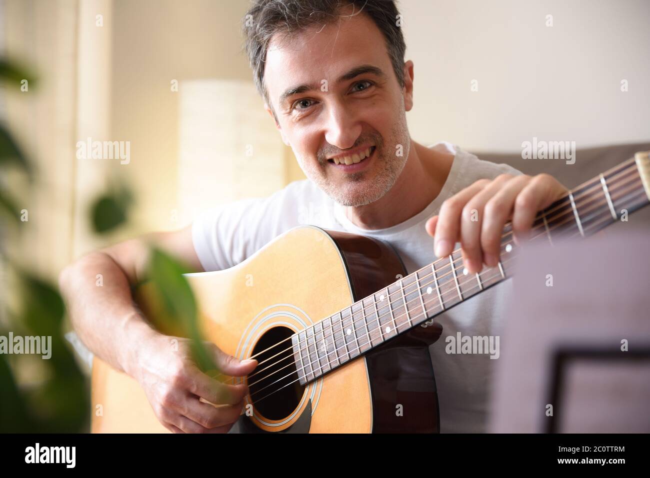 Lächelnder Mann mit seiner akustischen Gitarre in der Hand und Augen auf dem Sofa sitzend mit Musikständer mit Songs zu Hause Detail Stockfoto