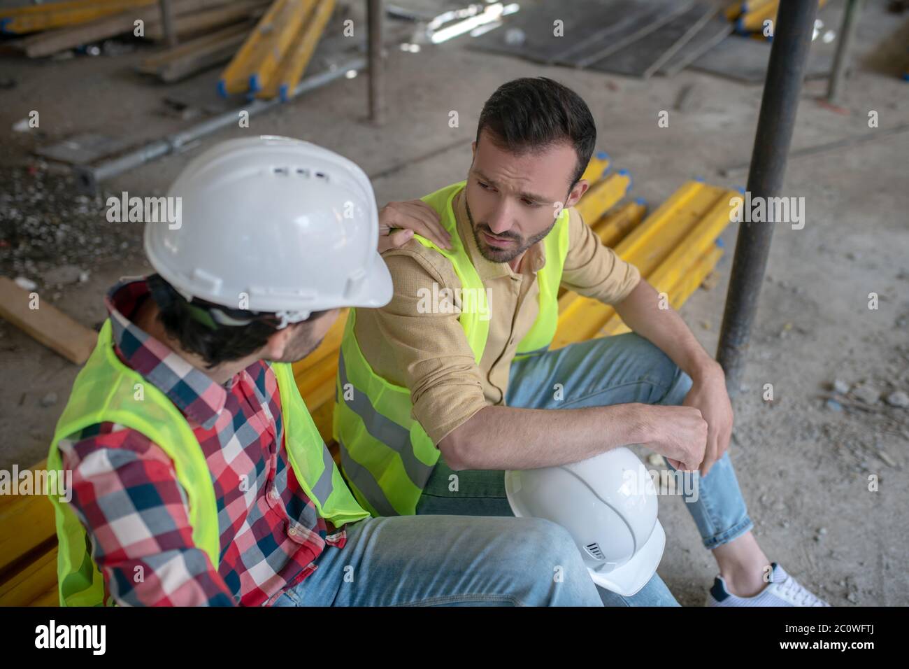 Müde Bauarbeiter in gelben Westen sitzen auf Brettern, einer von Männern legte seine Hand auf die Schulter des Kollegen Stockfoto