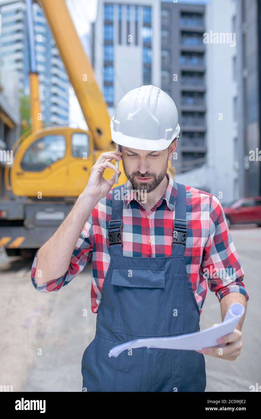 Bauarbeiter in Helm und kariertem Hemd vor dem Kran stehen, Blaupause halten, telefonieren Stockfoto