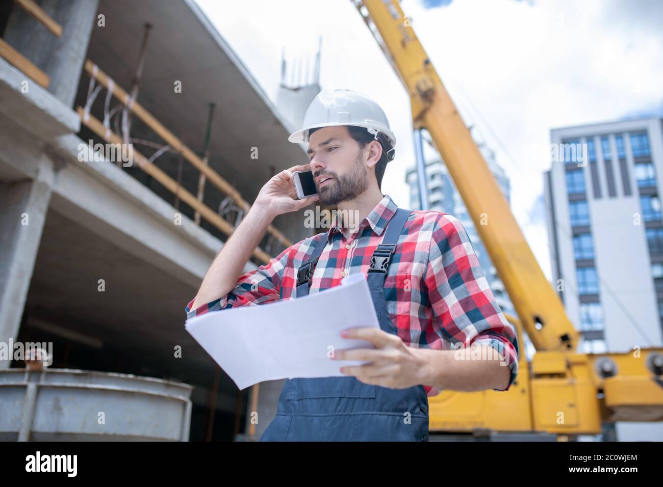 Bauarbeiter in Helm und kariertem Hemd halten Blaupause, Gespräch am Telefon Stockfoto
