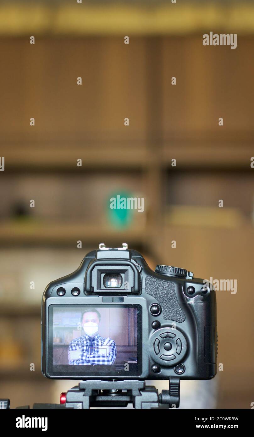 Ein Blogger in einer Schutzmaske fotografiert sich selbst vor der Kamera.  Der Fokus liegt auf seiner Kamera auf einem Stativ und dem Bild darauf.  Alles andere ist verschwommen Stockfotografie - Alamy