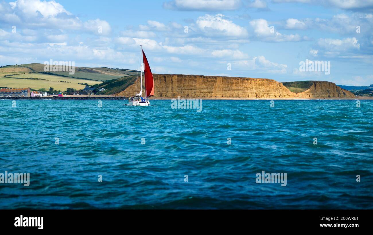 Eine Segelyacht passiert das Sandstein East Cliff, an der Jurassic Coast in der Nähe von West Bay, Dorset, UK. Berühmt gemacht durch die TV-Serie Broadchurch Stockfoto