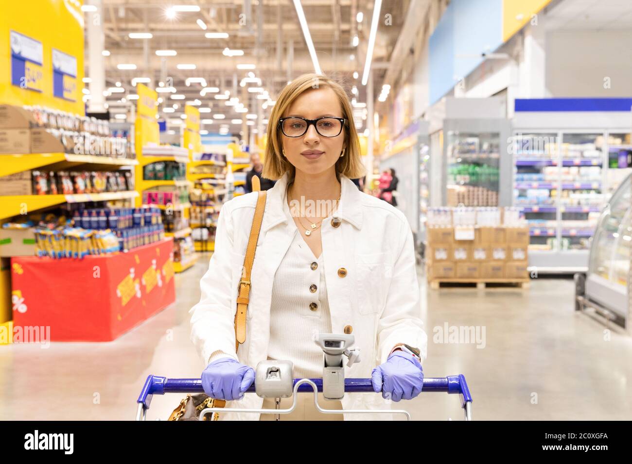 Kaukasische Frau, die im Supermarkt mit Einkaufswagen mit Waren zu Fuß, trägt Schutzhandschuhe aus Gummi. Ausbruch des Coronavirus. Stockfoto
