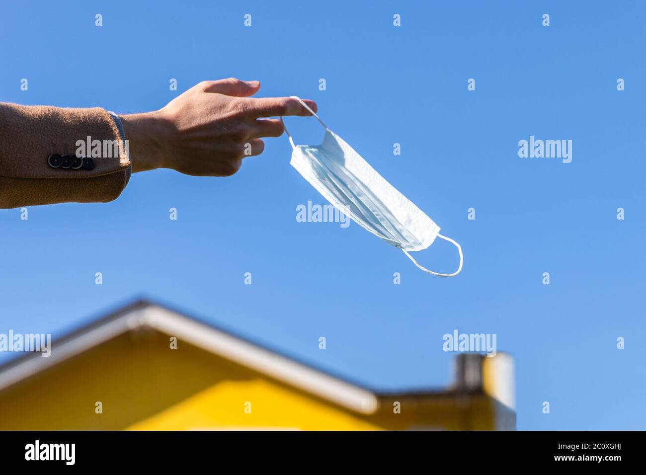 Mann zieht medizinische Schutzmaske aus und hält sie am Finger am blauen Himmel und gelbes Haus auf dem Hintergrund, genießt das Leben, saubere frische Luft nach Covid- Stockfoto
