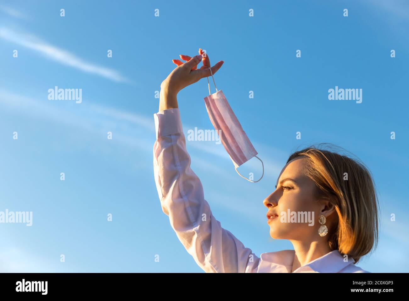 Glückliche Frau zieht medizinische Schutzmaske hält es auf ihrem Finger auf blauem Himmel Hintergrund, genießt das Leben, saubere frische Luft nach Covid-19 Pandemie, sel Stockfoto