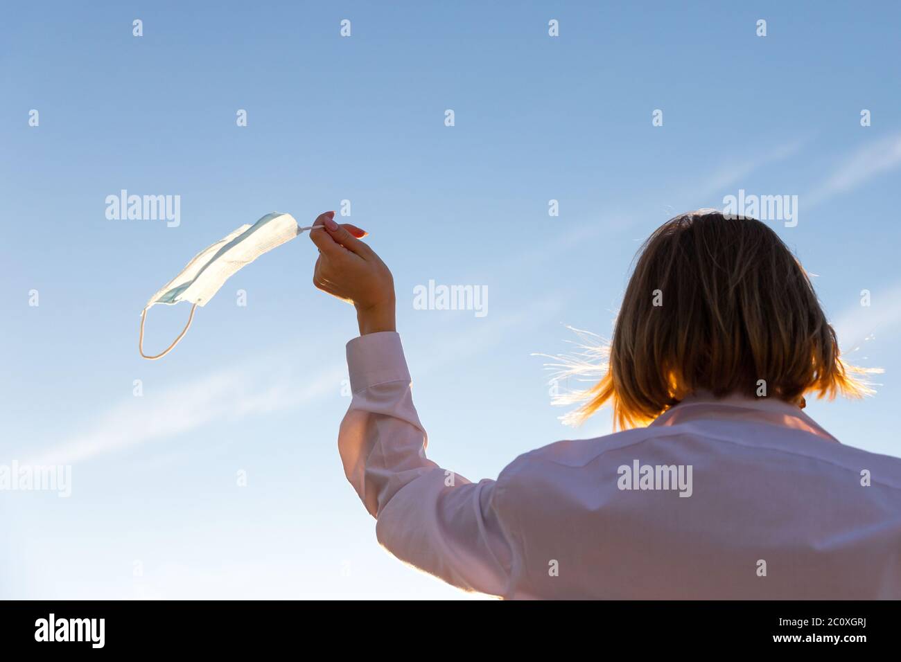 Glückliche Frau zieht medizinische Schutzmaske hält es auf ihrem Finger auf blauem Himmel Hintergrund, genießt das Leben, saubere frische Luft nach Covid-19 Pandemie, sel Stockfoto