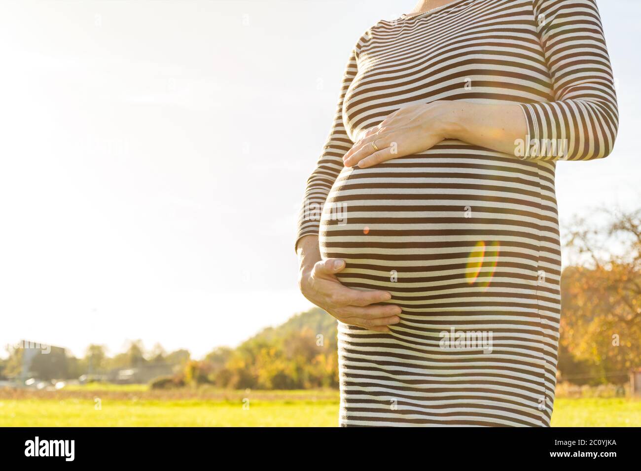 Frau, die auf der Wiese steht und ihren schwangeren Bauch im schönen Nachmittagslicht hält Stockfoto