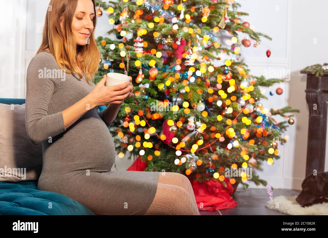 Schwangere Frau trinken heißen Tee und sitzen auf dem Sofa in der Nähe des Neujahrsweihnachtbaums Stockfoto
