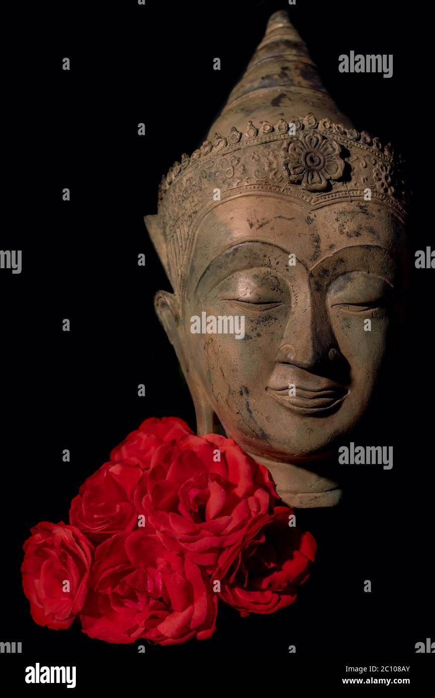 Traditionelle buddhistische Kopfstatue mit roten Rosen. Zen-Buddhismus, Achtsamkeit und Liebe. Gesicht des achtsamen Buddha in ruhiger Meditation. Spirituelle Erleuchter Stockfoto