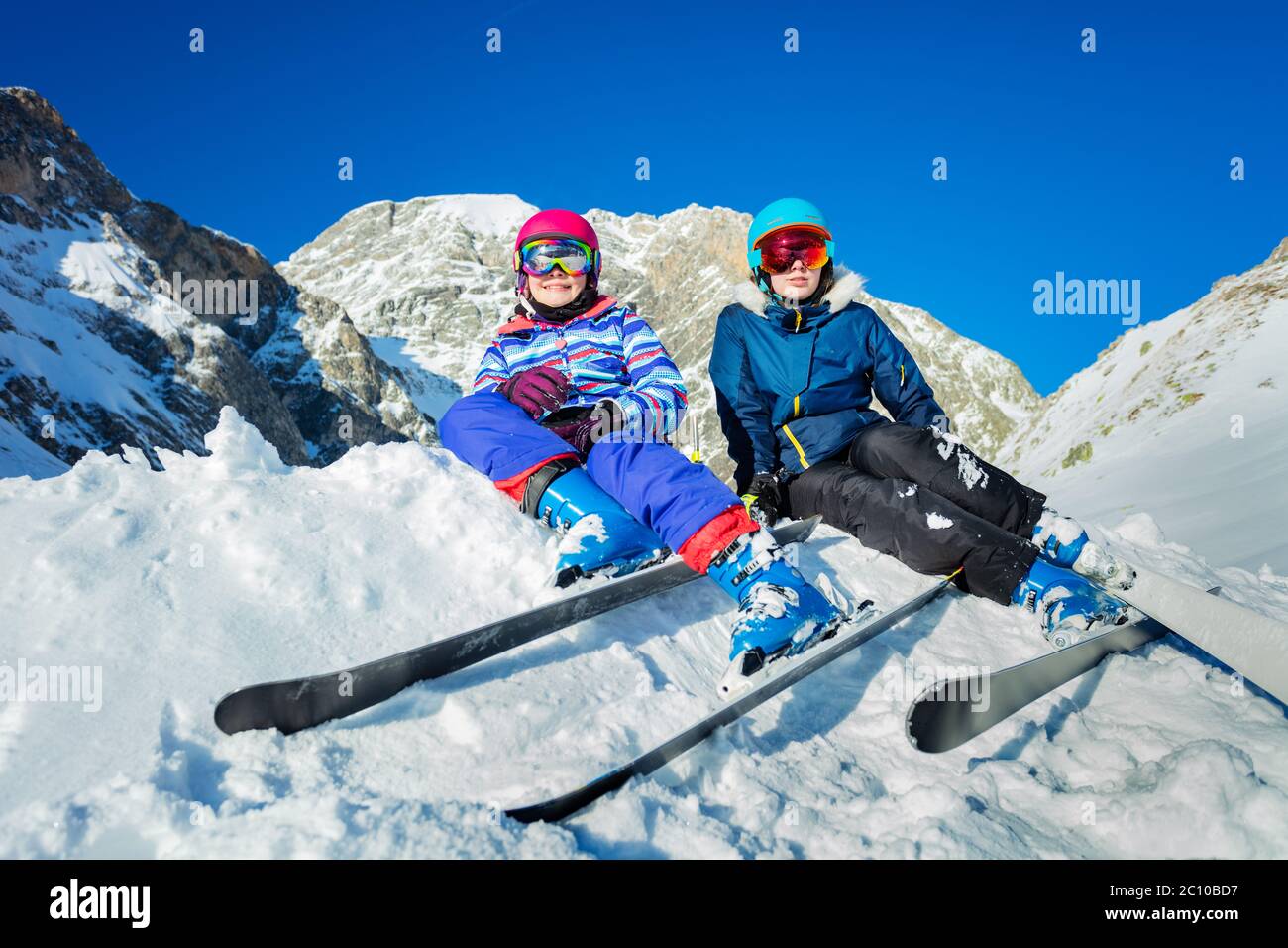 Zwei lächelnde glückliche Mädchen mit Bergski befestigt sitzen auf dem Haufen von Schnee in bunten Sport-Outfit Helm und Masken Stockfoto
