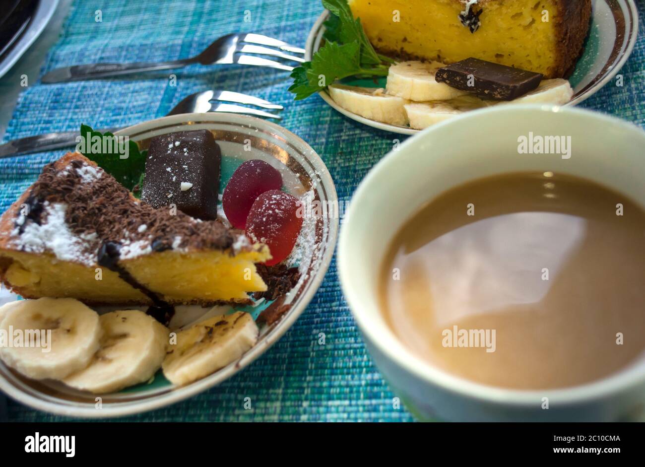 Portion Kuchen mit Bonbons, die mit Minze, Toffee, Banane und Gelee Bonbons, Puderzucker und einer Tasse Kaffee mit Milch auf einer blauen Tischdecke dekoriert sind Stockfoto