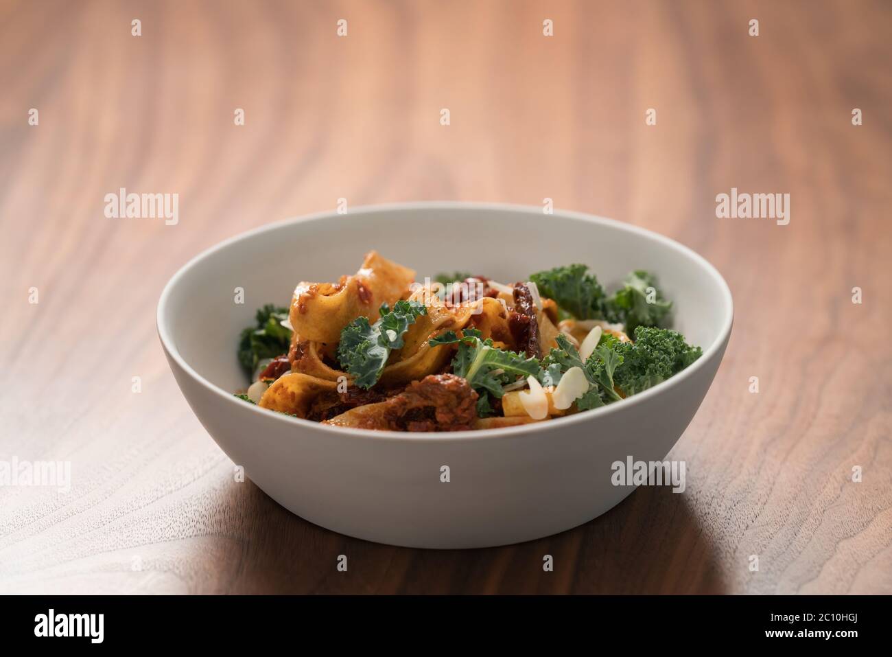 Fettuccine Pasta mit sonnengetrockneten Tomaten, Mandelflocken und Grünkohl in weißer Schale auf Walnusstisch Stockfoto