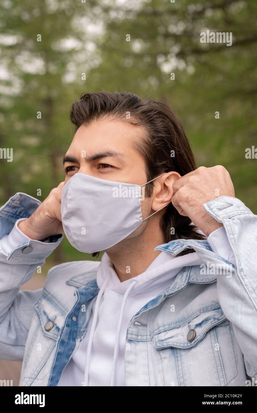 Brunette Mann in Denim Jacke im Freien stehen und setzen Stoffmaske auf Gesicht Stockfoto