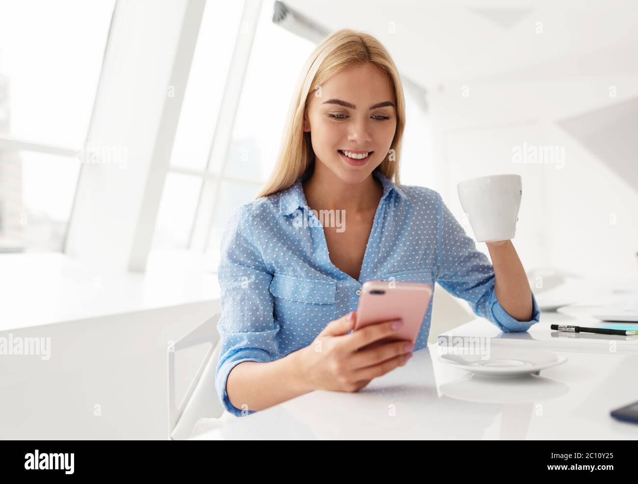 Lächelnde junge Frau mit Handy in der Hand, während sie Kaffee in einem weißen Raum trinkt Stockfoto