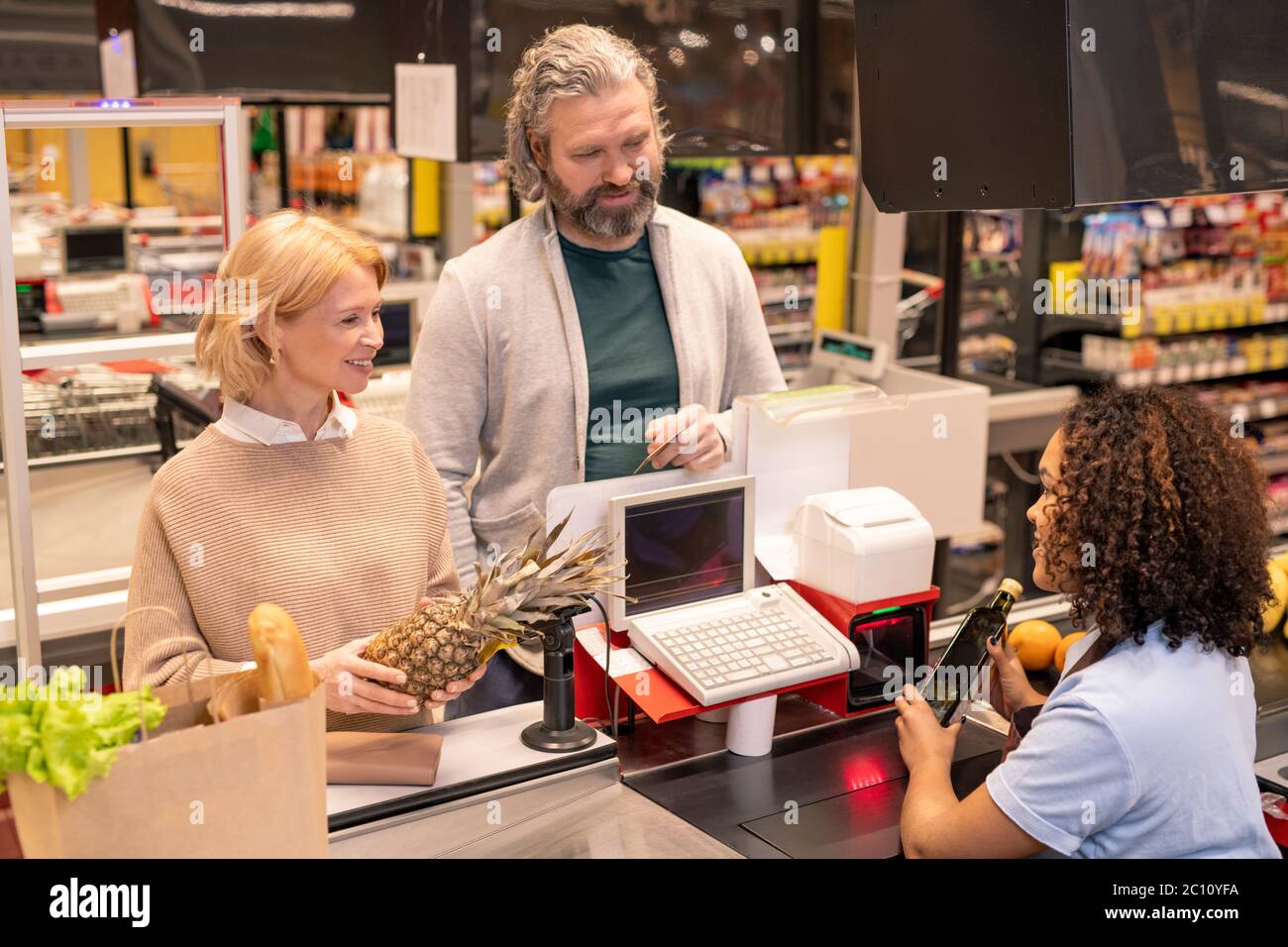 Reifes Paar steht an der Kasse im Supermarkt, während junge Mischrasse Frau Scannen Lebensmittel, die sie gekauft Stockfoto