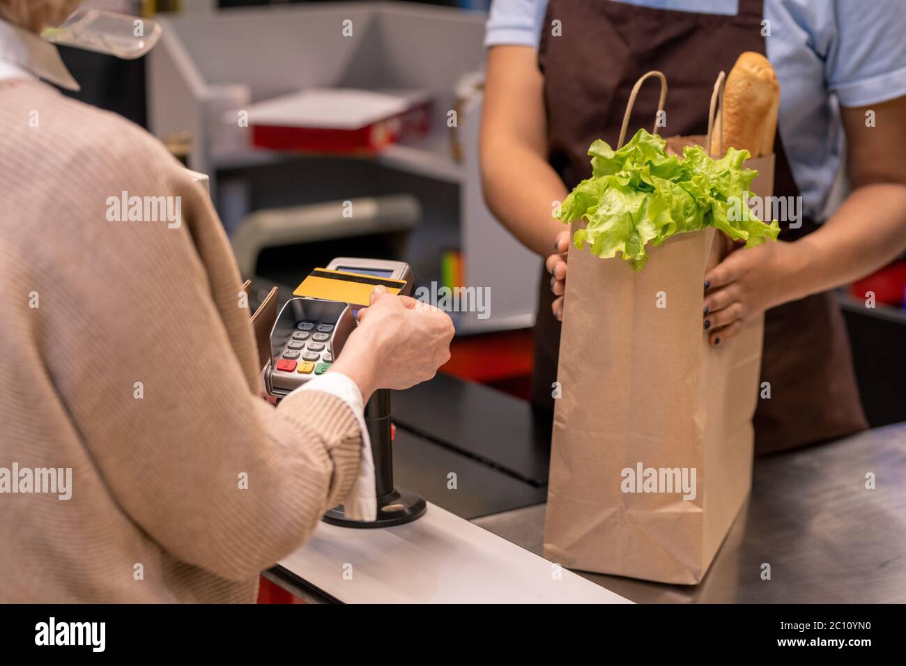 Hand der reifen weiblichen Kunden hält Plastikkarte über Bildschirm des Zahlautomaten durch Kassiererschalter beim Bezahlen für Lebensmittelprodukte Stockfoto