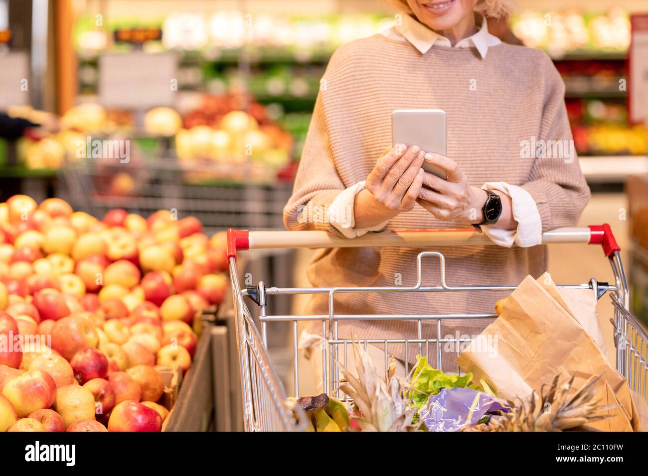 Reife weibliche Verbraucher in gestrickten beige Pullover schieben Warenkorb mit Lebensmitteln in Supermarker und Scrollen in Smartphone Stockfoto