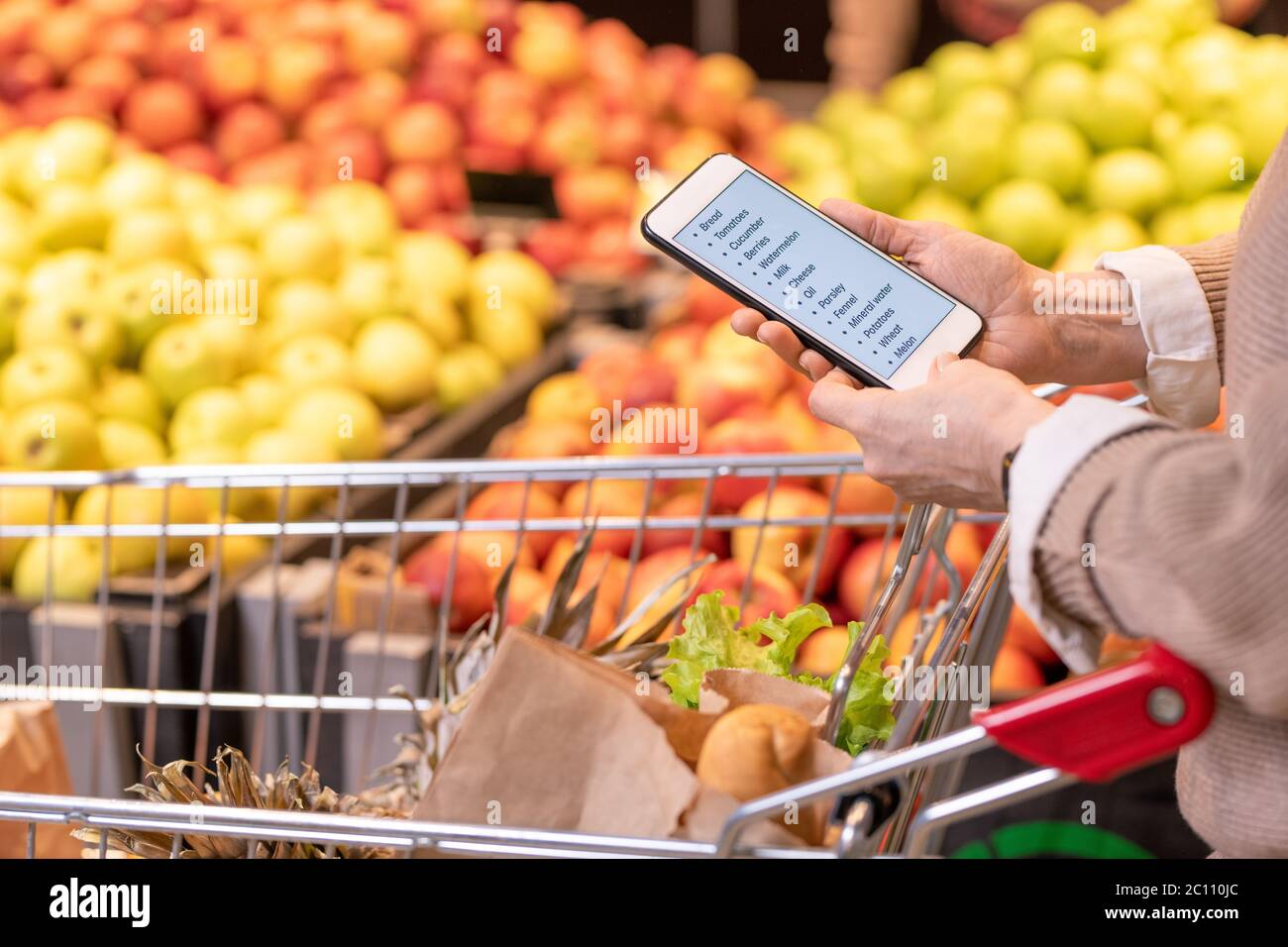 Hände von modernen reifen weiblichen Kunden mit Smartphone über Lebensmittel im Warenkorb Blick durch die Einkaufsliste Stockfoto