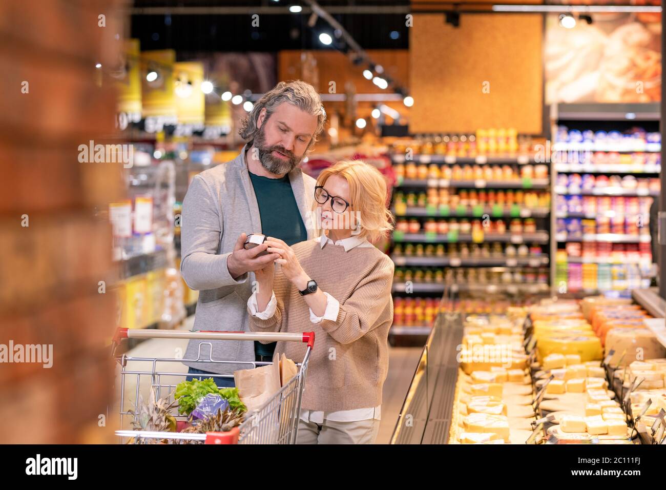 Reife blonde Frau zeigt ihren Mann Käse verpackt in runden Box, während beide die Auswahl Milchprodukte durch Display im Supermarkt Stockfoto