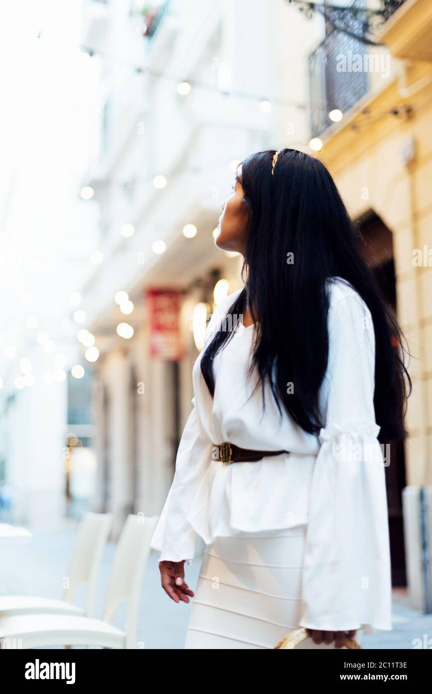 Latina Frau mit langen schwarzen Haaren und weißem Kleid drehte Blick auf eine Straße mit hängenden Lichtern. Urban Tourism Konzept Stockfoto