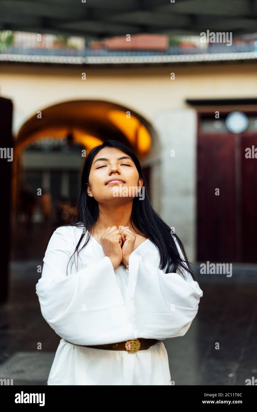 Latina Frau stehend mit langen schwarzen Haaren und weißem Kleid mit geschlossenen Händen und geschlossenen Augen in Meditationsposition, Friedenskonzept Stockfoto