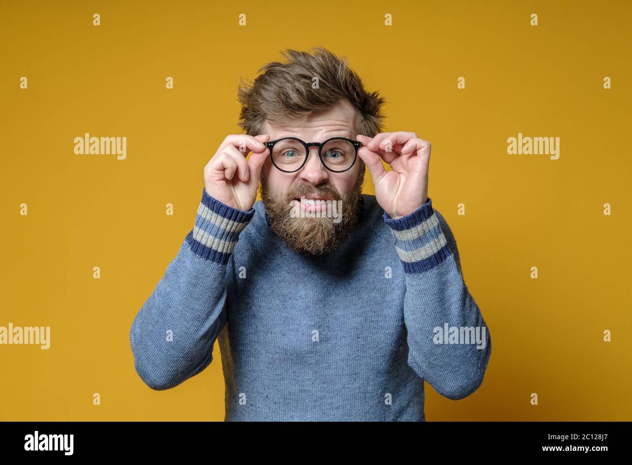 Der zottelige, bärtige Mann in einem alten Pullover mit einem seltsamen Gesichtsausdruck hält eine Brille in den Händen und versucht, etwas zu sehen. Stockfoto